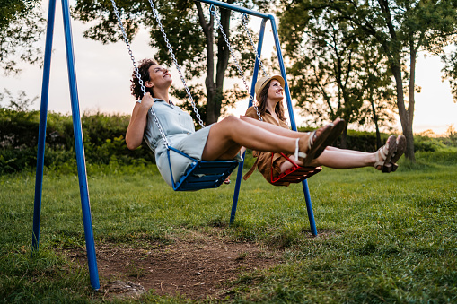 Two young smiling women riding swings in public park.
