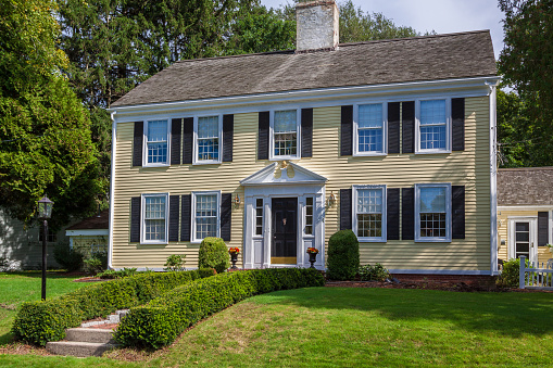 Luxury New England Colonial House with Yellow Clapboard Exterior, Grey Roof and Landscaped front yard with green grass and bushes,