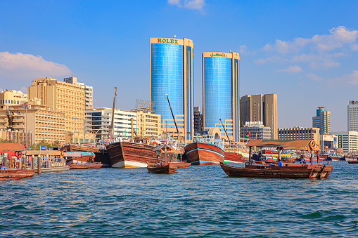 Dubai, United Arab Emirates - Abra boats move to and from the Abra station piers on the Deira side of Dubai Creek. In the background are Arab dhows moored at the quayside on Deira. The dhows are being unloaded and loaded for their next voyage. In the far background are the waterfront buildings and towers. Image shot in the afternoon sunlight from a boat on the Creek; horizontal format. Copy space.