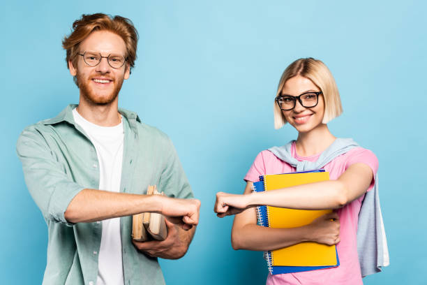 young students in glasses holding notebooks and books while fist bumping on blue