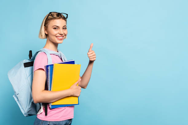 istock blonde student with backpack holding notebooks and showing thumb up on blue 1270533048
