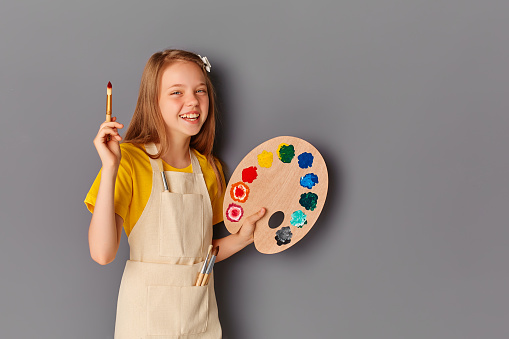 Cute happy teen girl with paint palette and brush on grey background