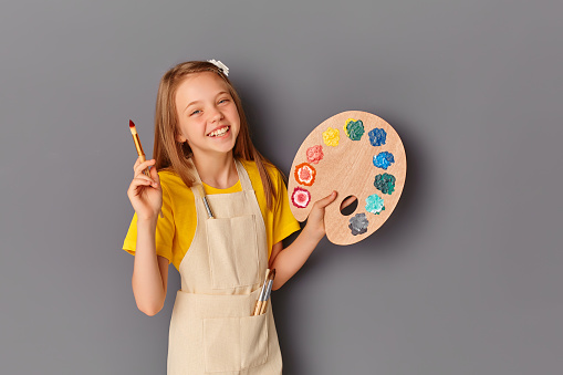 Portrait of a beautiful young teenage girl holding a wooden palette and brush against a gray wall. A smiling child with an art palette and an artist's brush in his hands. Items for children's creativity..