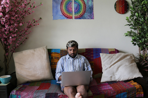 Stock photo showing an asian man sitting on lounge sofa covered with a brightly, multicoloured patchwork quilt and patterned scatter cushions. The man has his legs stretched out in front of him and is working on a laptop. Working from home concept.