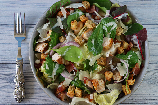 Stock photo showing a close-up, elevated view of healthy eating image of a salad bowl containing a chicken caesar salad with lettuce, red onion, beetroot leaves, cubed bacon, roasted chicken, topped with croutons and shavings of parmesan cheese.