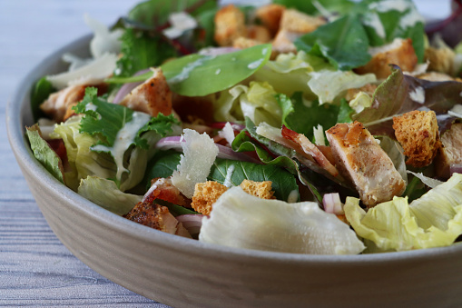 Stock photo showing a close-up of healthy eating image of a salad bowl containing a chicken caesar salad with lettuce, red onion, beetroot leaves, cubed bacon, roasted chicken, topped with croutons and shavings of parmesan cheese.