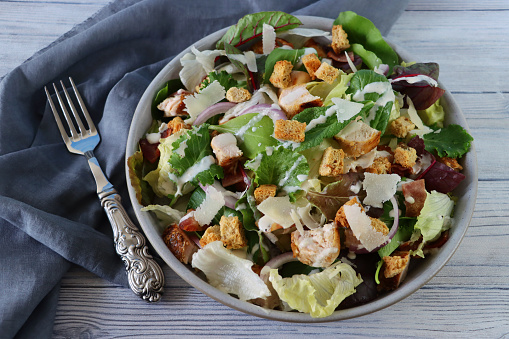 Stock photo showing a close-up, elevated view of healthy eating image of a salad bowl containing a chicken caesar salad with lettuce, red onion, beetroot leaves, cubed bacon, roasted chicken, topped with croutons and shavings of parmesan cheese.