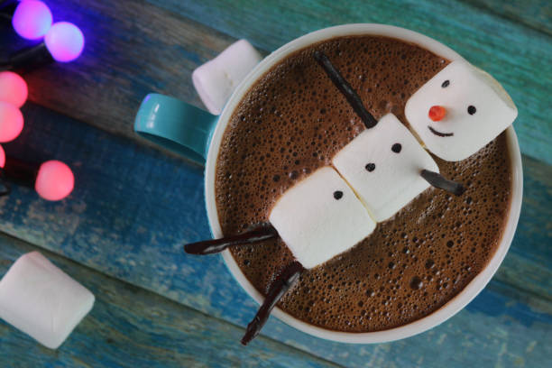 Image of marshmallow snowman sitting in turquoise blue mug of bubbly hot chocolate against a turquoise blue tongue and groove effect background, surrounded by fairy lights and marshmallows, elevated view Stock photo showing a turquoise blue mug of hot cocoa drink containing a snowman made from three marshmallows against a Christmas backdrop. froth decoration stock pictures, royalty-free photos & images