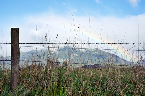 Looking through a Barbed Wire Fence to a Rural Landscape with Distant Rainbow.