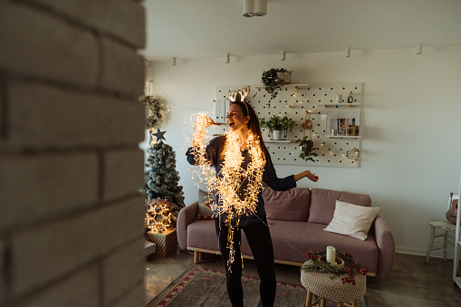 Photo of an excited young woman singing while decorating the living room of her apartment; wrapped in Christmas lights and wearing shiny reindeer antlers; preparing for the upcoming holidays.