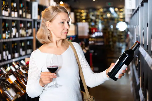 Portrait of glad  positive smiling mature woman tasting red wine while visiting winehouse in search of bottle of good wine