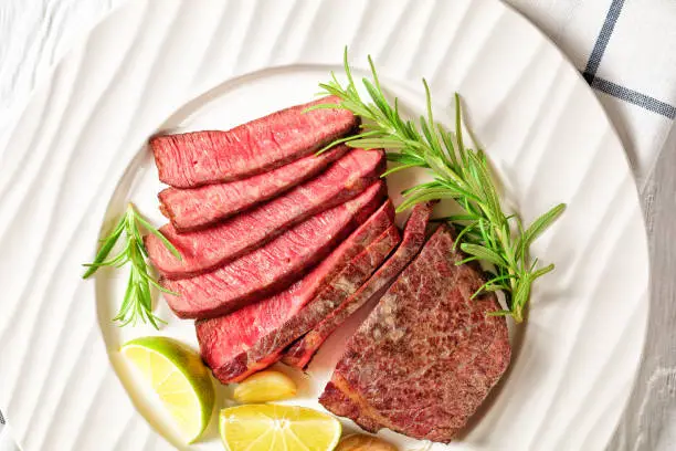 Medium rare steak beef striploin pan-seared and sliced, served on a white plate with rosemary and lime wedges on a white wooden table, , top view, close-up