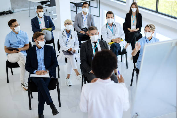 Large group of business people and healthcare workers with face masks on educational seminar. High angle view of group of healthcare workers and business people wearing protective face masks while attending a seminar at conference hall. attending photos stock pictures, royalty-free photos & images