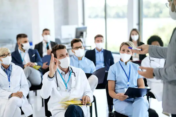 Photo of Happy doctor with protective face mask raising hand to ask a question while attending educational event.