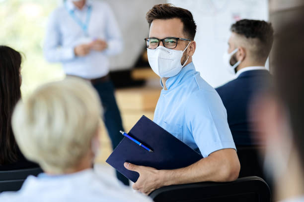 Happy entrepreneur wearing protective face mask while attending business seminar. Happy businessman with protective face mask attending an educational event and looking at camera. kn95 face mask photos stock pictures, royalty-free photos & images