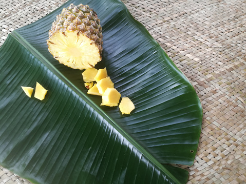 Pineapple and its slices placed in a green banana leaf.The cross section of a full pineapple is visible.Its pieces are placed near to it.Placed in a green banana leaf.
