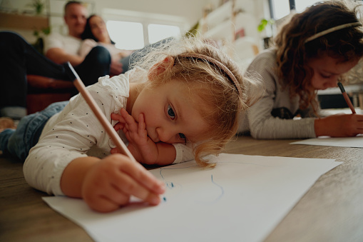 Family bonding time - little girl sitting with her sister drawing on the floor with her parents in the background