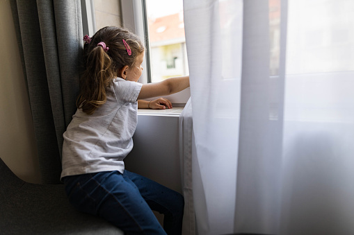 Small girl relaxing by the window at home and looking through it.