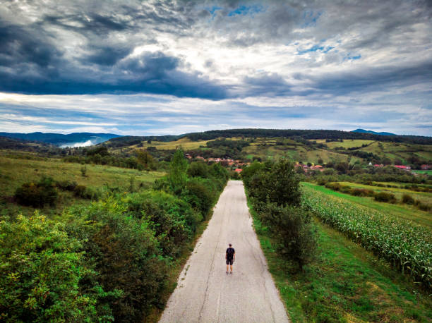Drone image depicting an aerial view of a backpacker walking alone in the middle of a straight rural country road, surrounded by agricultural meadows and forest. Room for copy space.