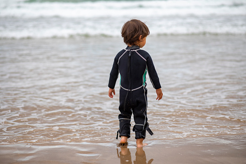 Rear view of a young mixed race child standing in a wet suit on the water's edge looking out to the sea.