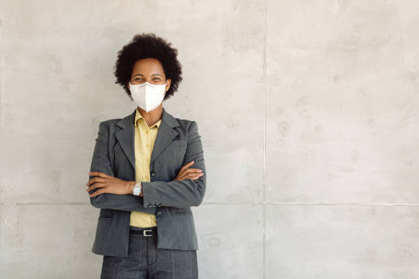 Confident black businesswoman standing with crossed arms while wearing protective face mask. Happy African American businesswoman with face mask standing against the wall and looking at camera. Copy space. kn95 face mask photos stock pictures, royalty-free photos & images