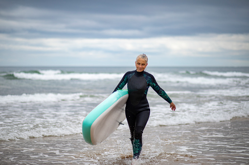 Wide shot of a mature woman walking out of the sea holding her surfboard. She is wearing a wetsuit.