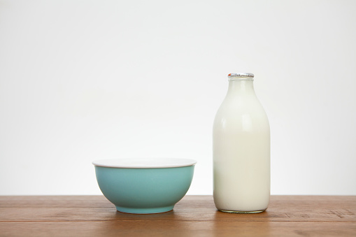 Single blue coloured cereal bowl next to a full pint of milk on a wooden table against a light coloured wall. Part of a series.