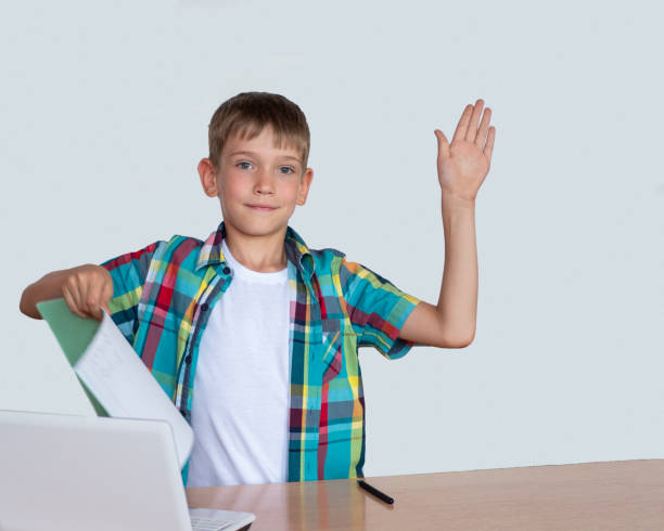 istock A smart, cute boy raised his hand, holds in his other hand a notebook with completed homework, a laptop on the table 1270602314