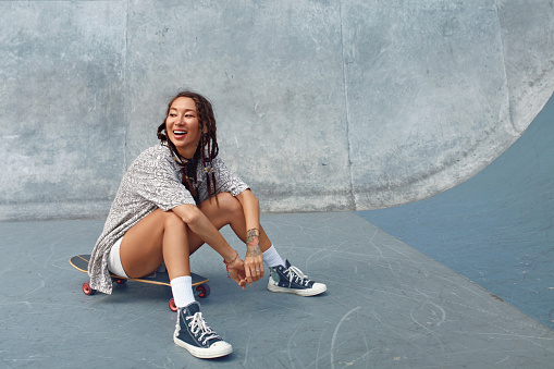 Retrato de Skater Girl en Skatepark. Adolescente femenina en traje casual sentado en el monopatín contra la pared de hormigón. Patinaje de verano con equipamiento deportivo moderno como parte del estilo de vida activo. photo