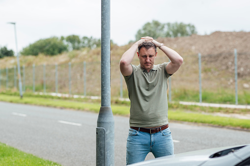 A caucasian male in his 30's standing at the side of the road after he has crashed into a lamp post.
