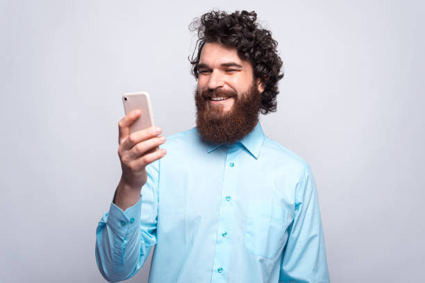 istock Cheerful young bearded hipster man in blue shirt using his smartphone and smiling 1270616925
