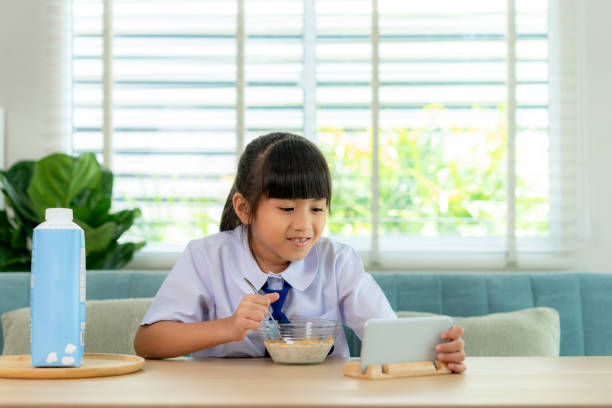 Asian elementary school student girl in uniform eating breakfast cereals with milk and looking cartoon in smartphone to distractions to eat it in morning school routine for day in life getting ready for school. estudiante de primaria asiática en uniforme comiendo cereales de desayuno con leche y buscando dibujos animados en el teléfono inteligente a distracciones para comerlo en la rutina de la escuela matutina para el día en la vida preparándose para la escu