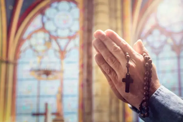 Hands folded in prayer in church with rosary beads and religious cross concept for faith, spirtuality and religion