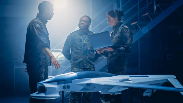 istock Army Aerospace Engineers Work On Unmanned Aerial Vehicle / Drone. Uniformed Aviation Experts Talk, Using Laptop. Industrial Facility with Aircraft for Performing Surveillance, Warfare Tactics, Attack 1270623266