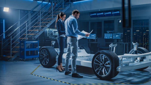 istock Auto Industry Design Facility: Male Chief Engineer Shows Car Blueprints Female Software Design and Integration Engineer. Electric Vehicle Platform Chassis Concept Has Wheels, Engine and Battery 1270623302