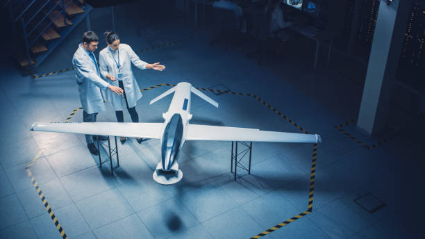 istock Two Aerospace Engineers Work On Unmanned Aerial Vehicle Drone Prototype. Aviation Scientists in White Coats Talking, Using Tablet Computer. Industrial Laboratory with Surveillance or Military Aircraft 1270623363