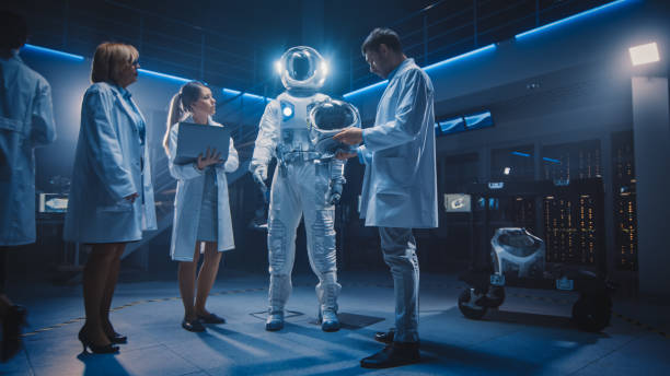 istock Team of Aerospace Scientists and Engineers Wearing White Coats have Discussion, Use Computers, Construct Astronaut Helmet for New Space Suit Adapted for Exploration and Travel. Low Angle Shot 1270623372