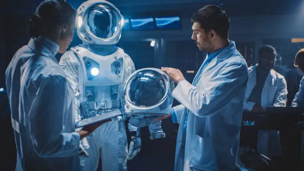 Photo of Diverse Team of Aerospace Scientists and Engineers Wearing White Coats have Discussion, Use Computers, Construct Astronaut Helmet for New Space Suit Adapted for Galaxy Exploration and Travel.