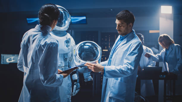 istock Diverse Team of Aerospace Scientists and Engineers Wearing White Coats have Discussion, Use Computers, Construct Astronaut Helmet for New Space Suit Adapted for Galaxy Exploration and Travel. 1270623536