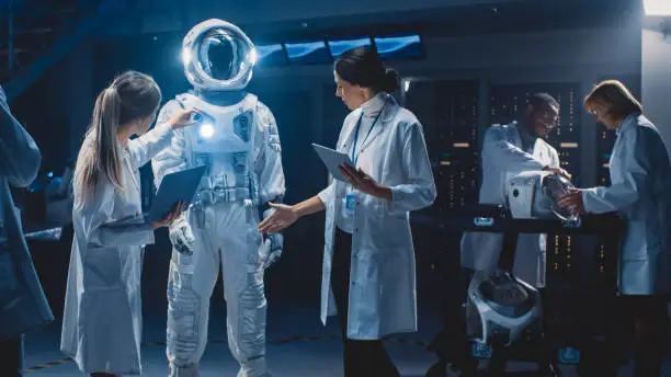 Photo of Diverse Team of Aerospace Scientists and Engineers Wearing White Coats have Discussion, Use Computers Design New Space Suit Adapted for Galaxy Exploration and Travel. Constructing Astronaut Suit