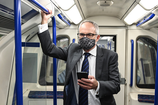 Front view of corporate professional in suit and homemade protective face mask holding grab bar while using smart phone on subway train in time of COVID-19.