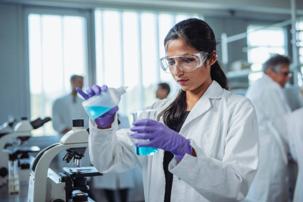 Indian University Student Pouring Blue Solution into Beaker Woman in early 20s wearing protective eyewear, white lab coat, and purple gloves pouring blue solution into beaker in educational lab. beaker pour stock pictures, royalty-free photos & images
