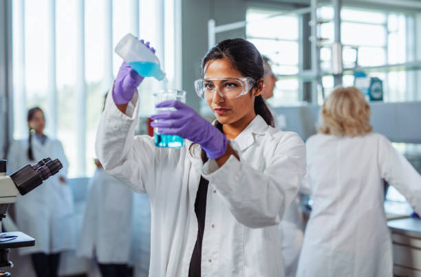 Female University Student Pouring Solution into Beaker Front view of Indian woman in early 20s wearing white lab coat, protective eyewear, and purple gloves smiling as she pours blue solution into beaker. beaker pour stock pictures, royalty-free photos & images