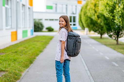Portrait of a cheerful caucasian schoolgirl standing by the school and wearing summer casual clothes and backpack. She is looking over shoulder at camera.