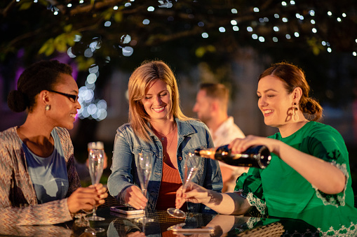 Group of diverse female friends drinking champagne outdoors at a bar/restaurant, celebrating and having fun together.