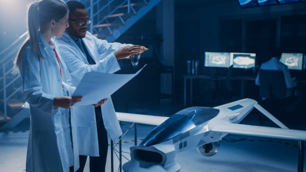 dos ingenieros aeroespaciales trabajan en vehículos aéreos no tripulados / prototipo de drones. científicos de aviación hablando, usando planos. laboratorio industrial con aeronaves de vigilancia aérea comercial - aerospace industry fotografías e imágenes de stock