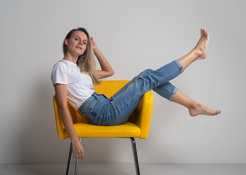 Beautiful young woman is sitting or lying on yellow chair with legs up and looking arrogantly at camera, studio shot
