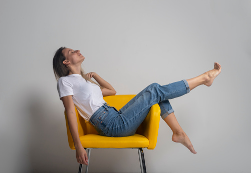 Beautiful young woman is sitting or lying on yellow chair with legs up and closing eyes with satisfaction and happiness, studio shot