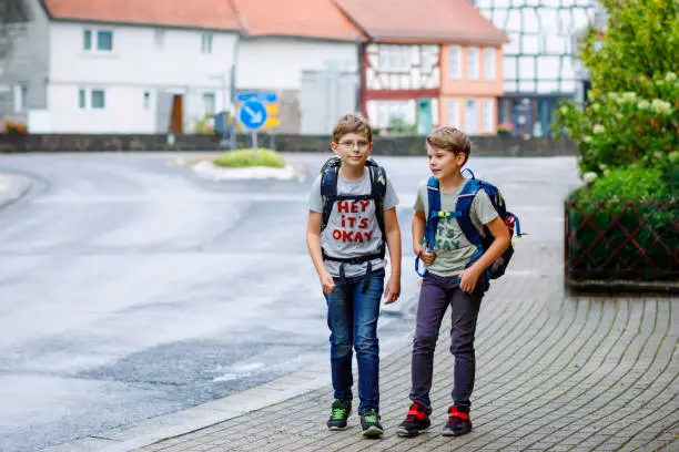Two little kid boys with backpack or satchel. Schoolkids on the way to school. Healthy adorable children, brothers and best friends outdoors on the street leaving home. Back to school. Happy siblings