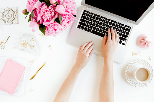 Female workspace with female hands, laptop, pink peonies bouquet, accessories on white background. Top view feminine background. Flat lay women's office desk. High quality photo